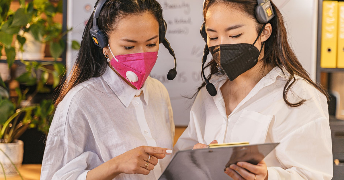 Two women in face masks and headsets discussing work on a clipboard in an office setting.